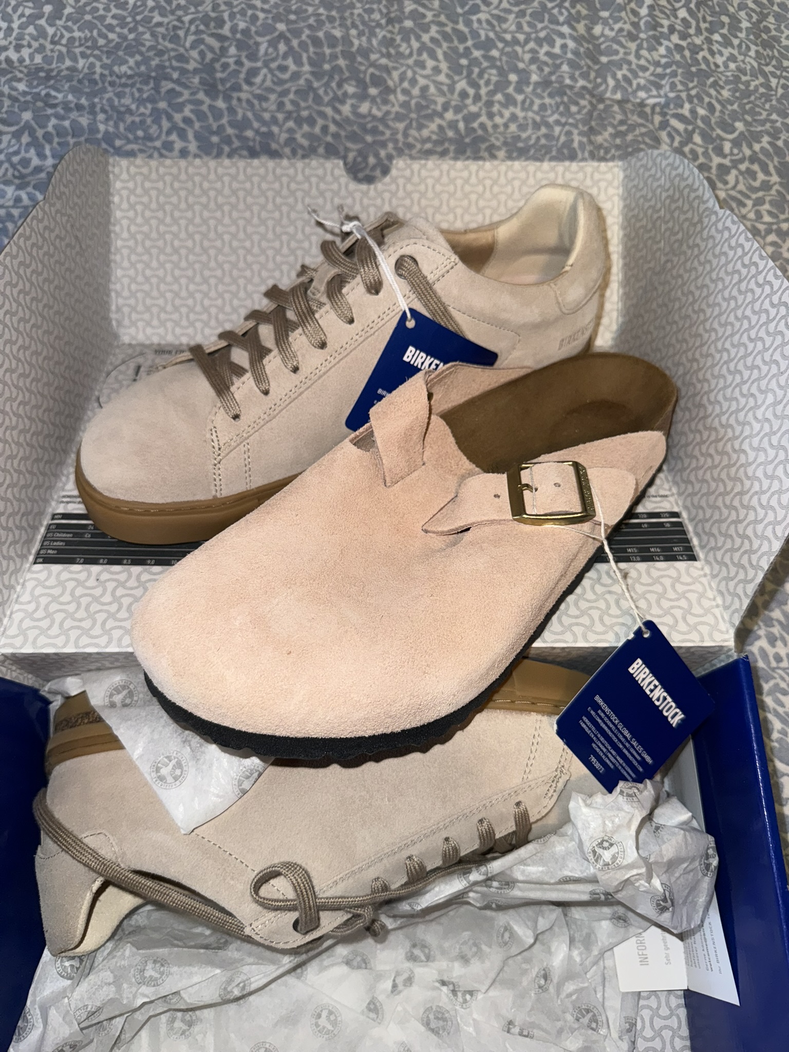A pair of tan suede Birkenstocks and sneakers unboxed with tags still on.