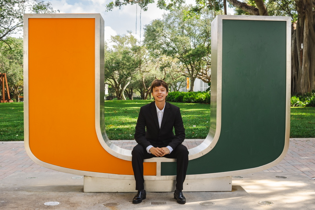 Bruno Veloso, smiling in a suit, seated on the University of Miami 'U' sculpture on campus.